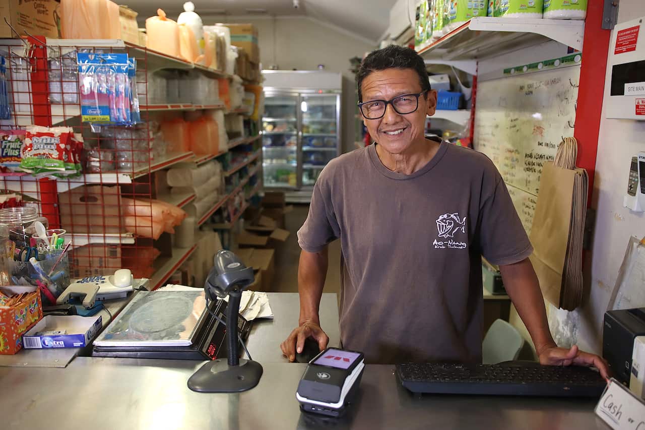 A shop owner wearing glasses and a T-shirt stands behind the counter at his convenience shop.