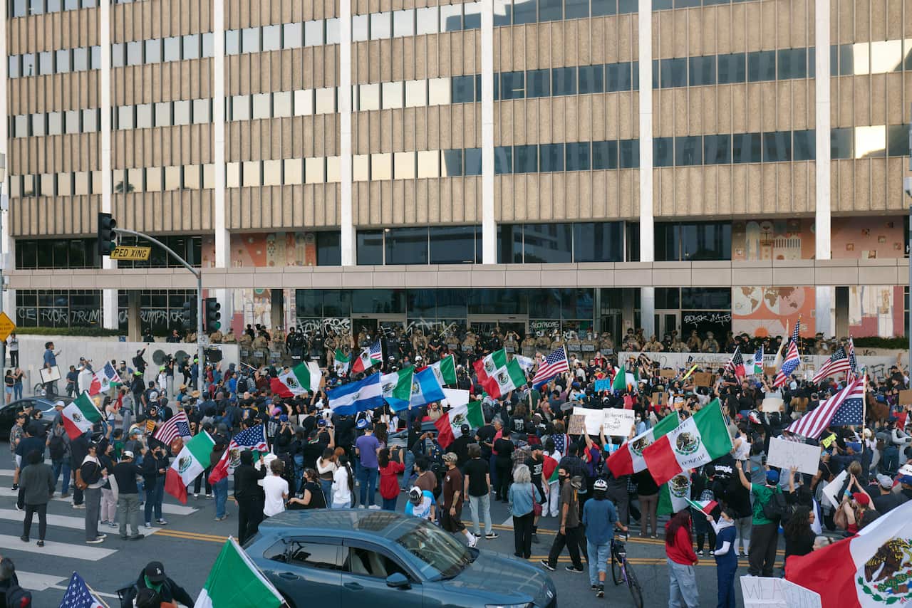 A large group of protesters, many holding Mexican flags, outside a building being guarded by troops.