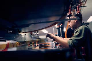 A man in profile carefully feeds a strip of IMAX film through a large, industrial projector mechanism inside a dimly lit projection booth.