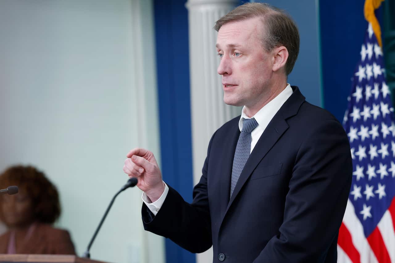 View of man in suit from side on. He stands in front of a podium, speaking into a microphone with his hand pointed. An American flag can be seen behind him.