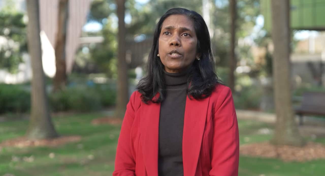 A woman wearing a black turtleneck and red blazer speaking in a park, near trees.