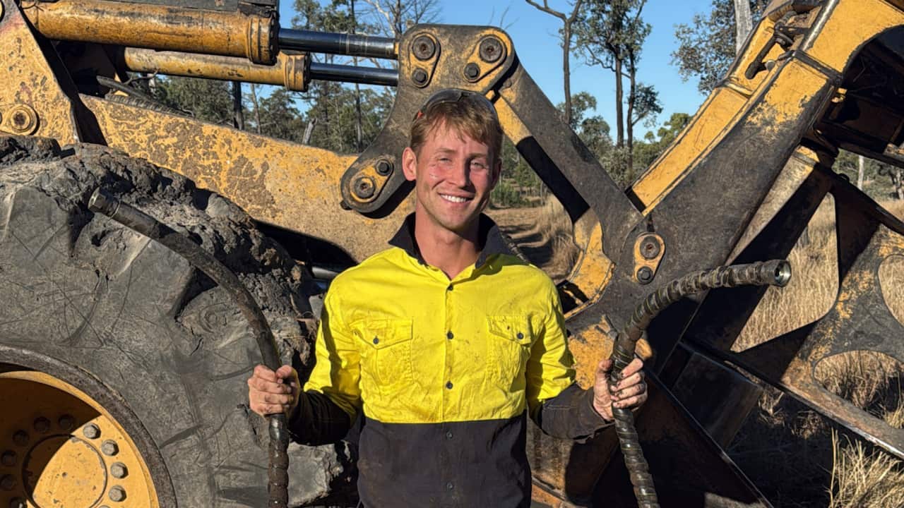 A smiling man wearing a yellow hi-vis shirt and holding a hose in each hand, standing in front of earth-moving machinery. 