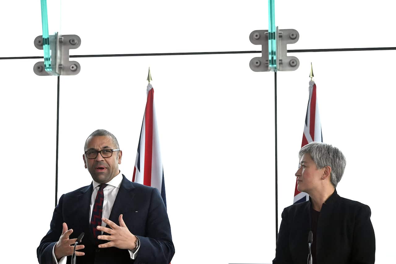 A man in a suit and glasses speaks at a lectern in front of flags, alongside a woman in a suit 