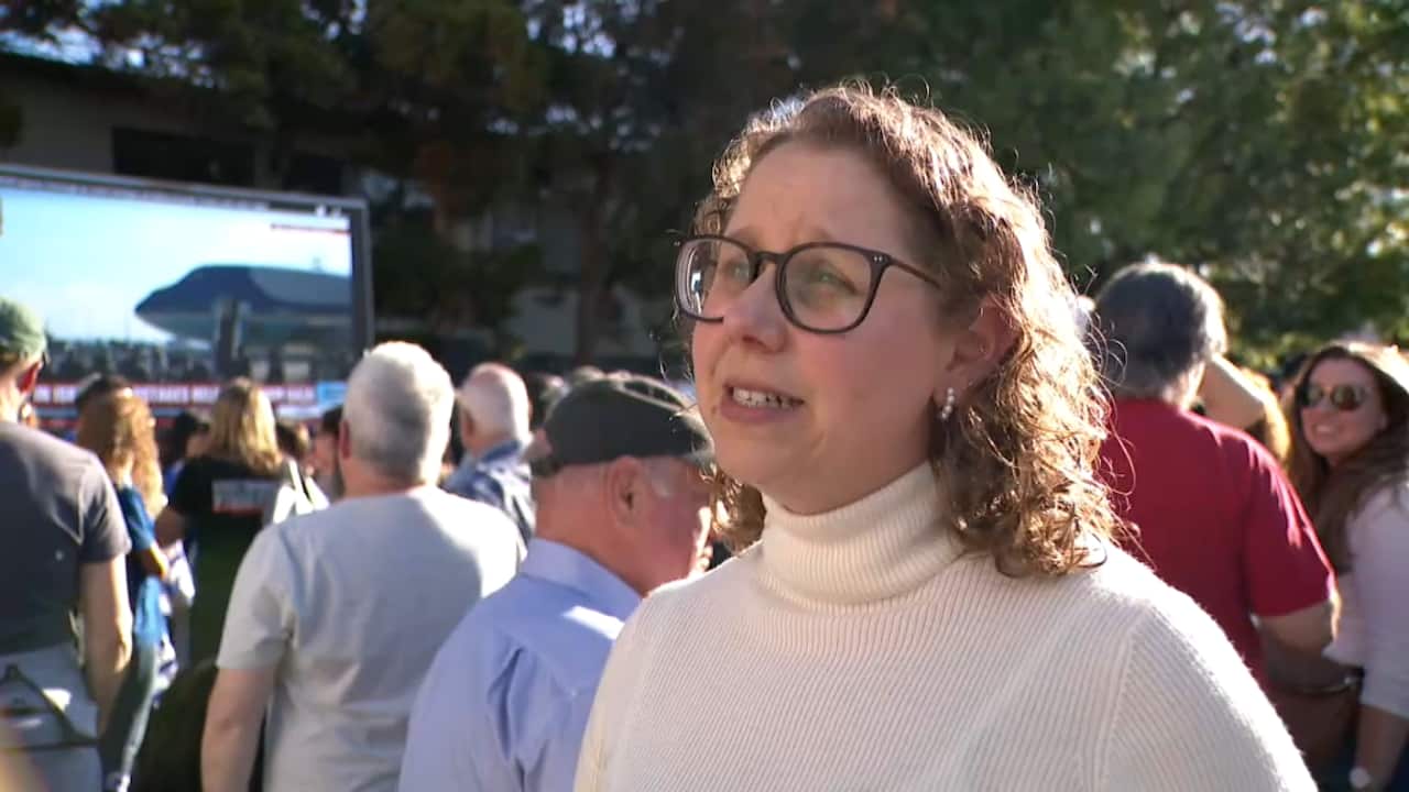A woman in a white turtle neck is looking sideways with a crowd visible behind her.