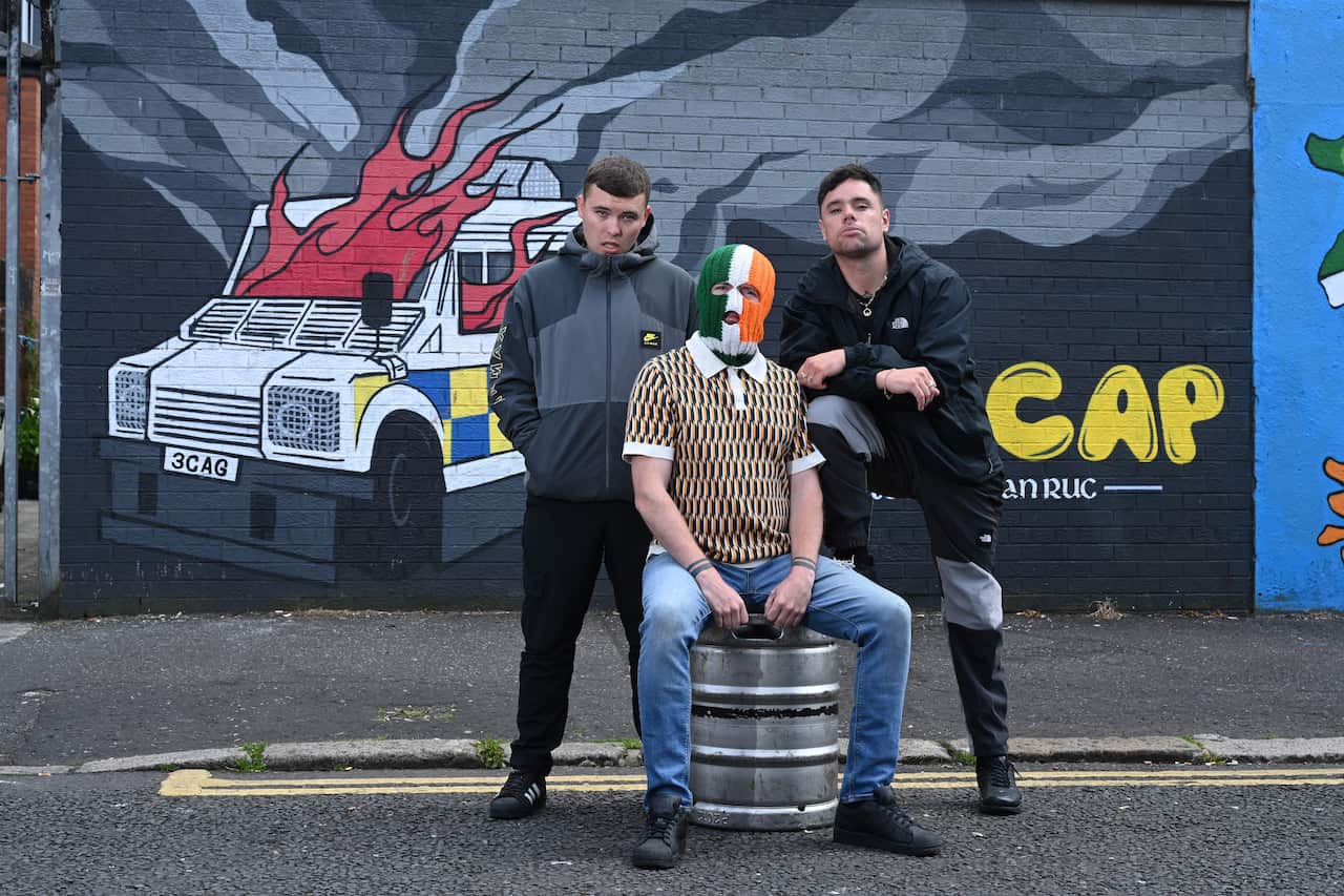 A man wearing a green, white and orange balaclava sitting outside on a steel beer keg. There is a man standing either side of him. They are in front of a mural featuring a drawing of an armoured white vehicle with flames coming from it.