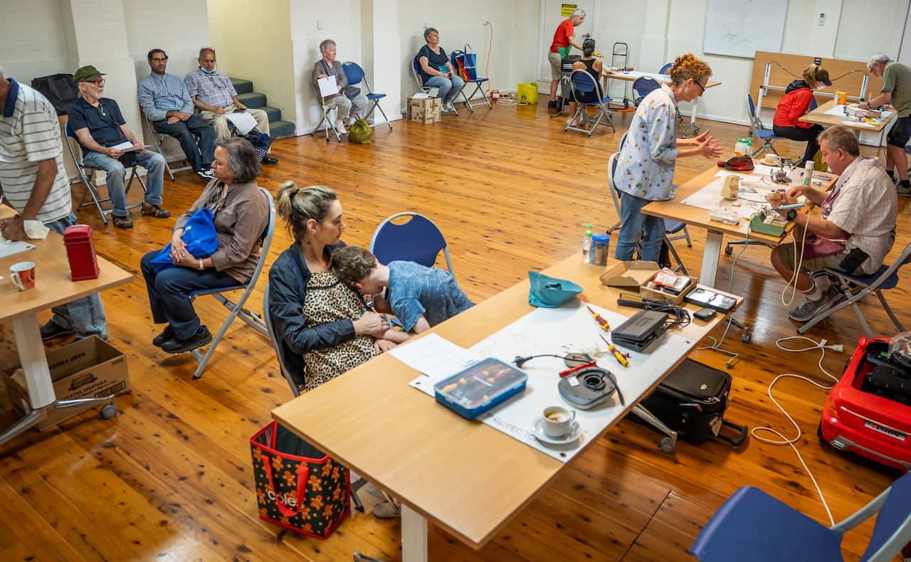 People sit on chairs in a large room with tables covered in electronic equipment.