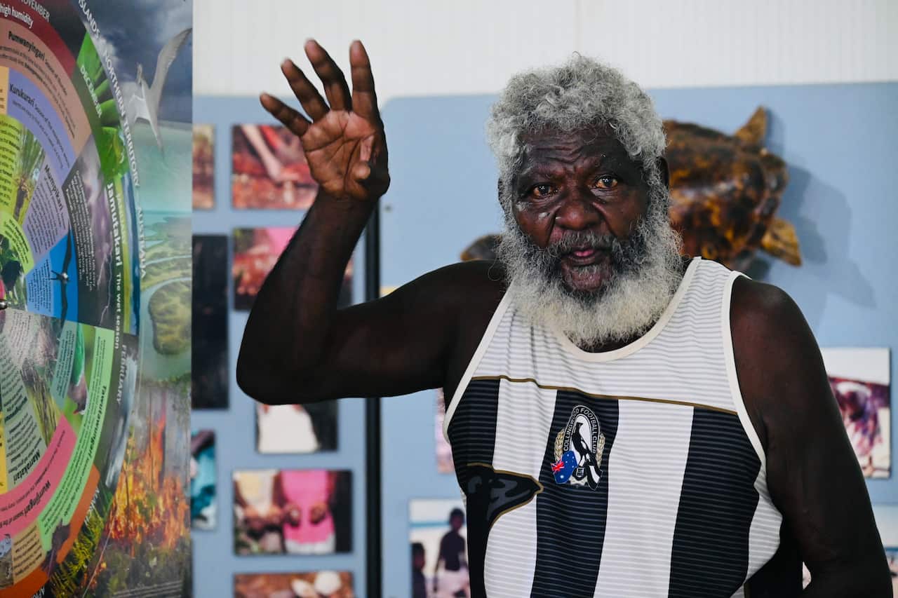 A man wearing a Collingwood Aussie Rules jumper speaks with his right hand raised towards a colourful poster