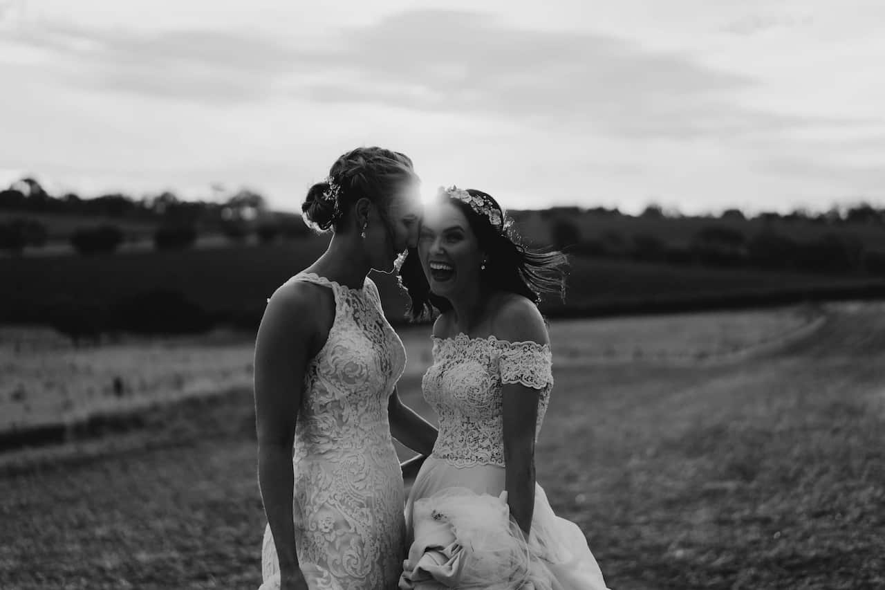 Black and white photo of two women on their wedding day. Both are wearing white wedding dresses and standing in the countryside.