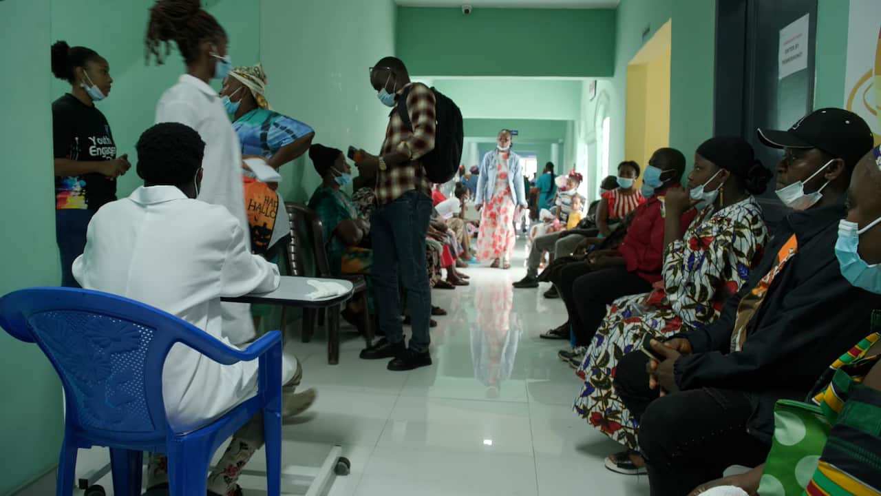 A crowded hospital waiting area with patients seated along both sides of a corridor. Several people are wearing face masks, and a staff member in a white coat sits at a desk in the foreground.