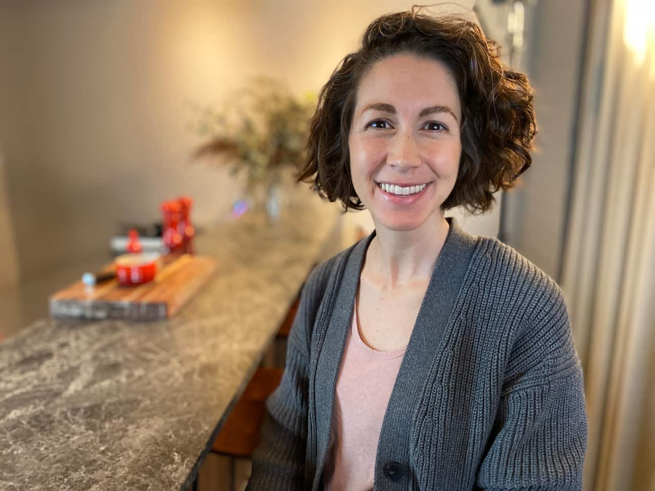 A woman wearing a grey pullover on a pale pink top, sitting indoors, smiles at the camera.