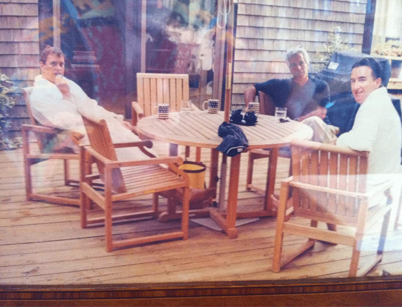 Three men, two of them wearing white bathrobes, and the other wearing a blue shirt site at an outdoor table on a deck. 