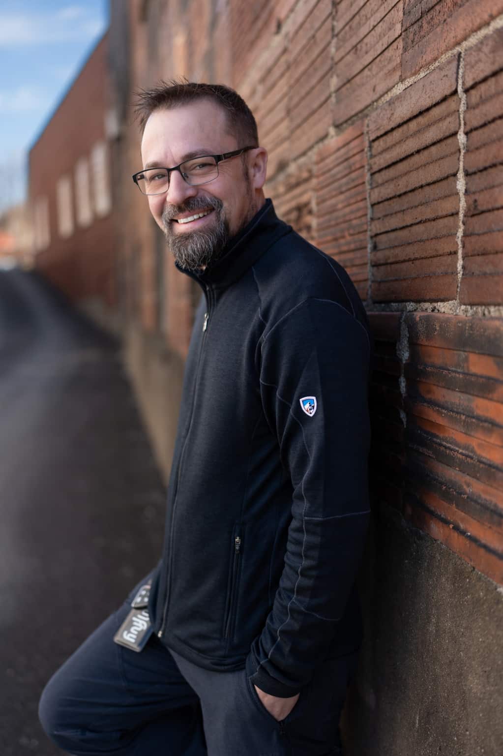 A middle-aged white man, leaning against an outdoor wall. He is dressed in black, with glasses, and looking to his left while smiling.