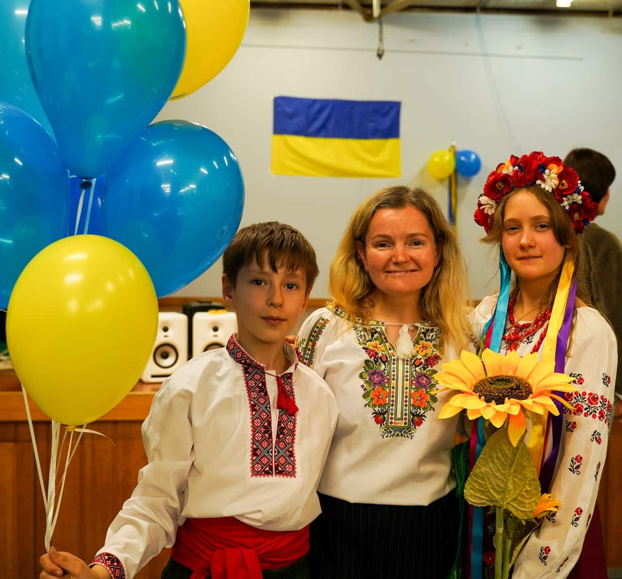 A woman and two children in traditional Ukrainian dress smile to the camera, yellow and blue balloons, the colours of the Ukrainian flag, are beside them.