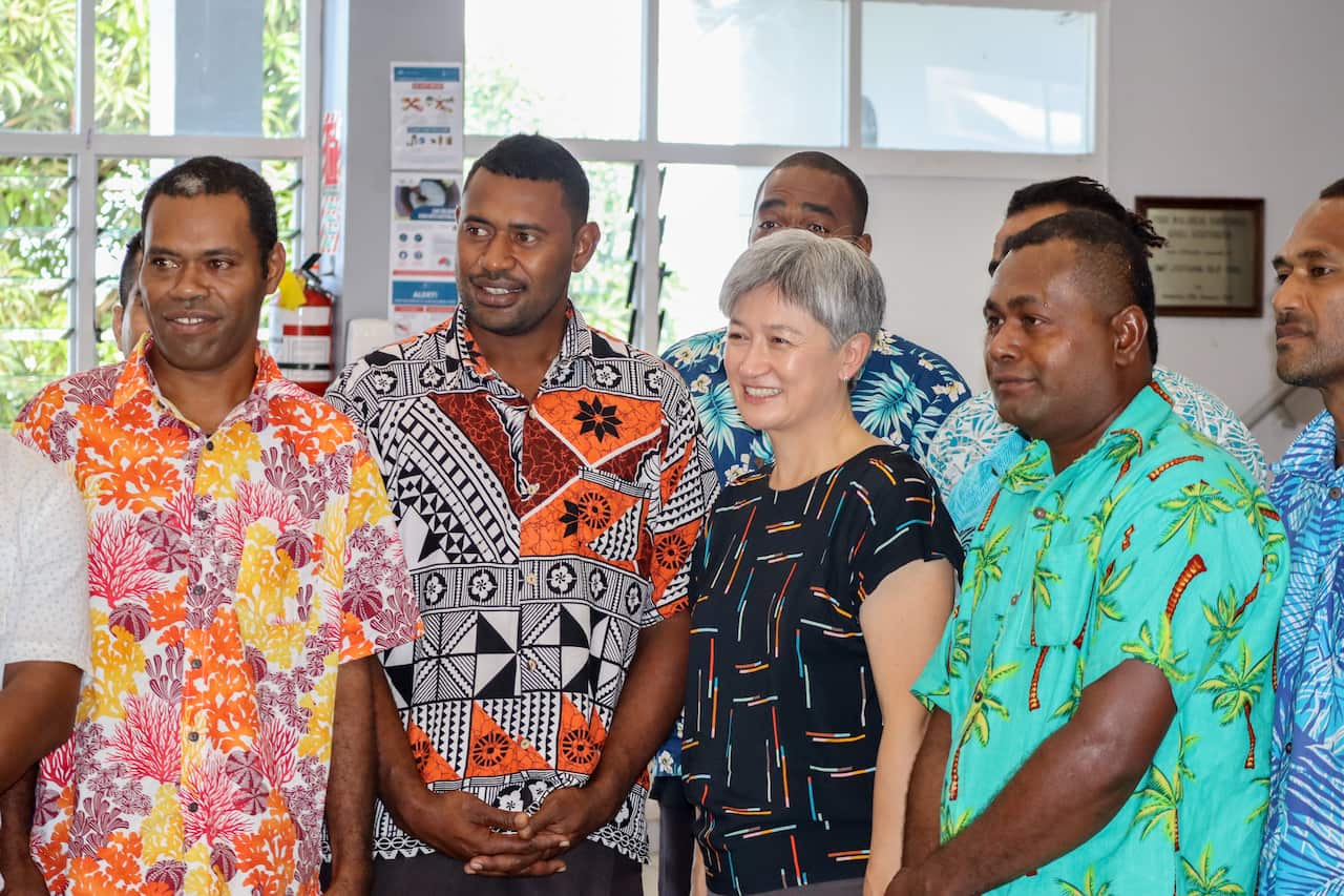 Penny Wong, wearing a patterned t-shirt, stands among a group of Fijian men who are wearing colourful patterned shirts and smiling.
