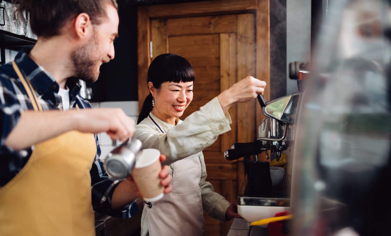 Two Smiling Colleagues Making Take Away Coffee Behind The Counter Of A Coffee Shop
