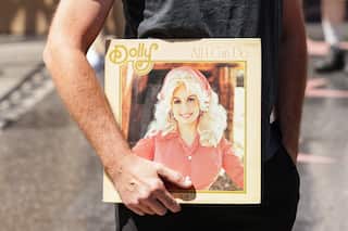 Person holding a vinyl record album with a portrait of Dolly Parton on the cover, standing outdoors on a paved walkway with the record tucked under one arm.