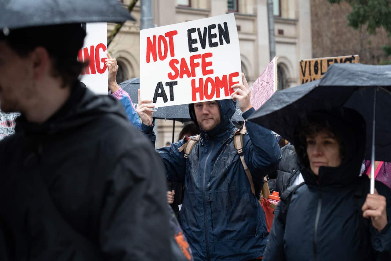 A crowd of people in black clothing carrying umbrellas in the rain. One man holds a sign saying, Not Even Safe at Home