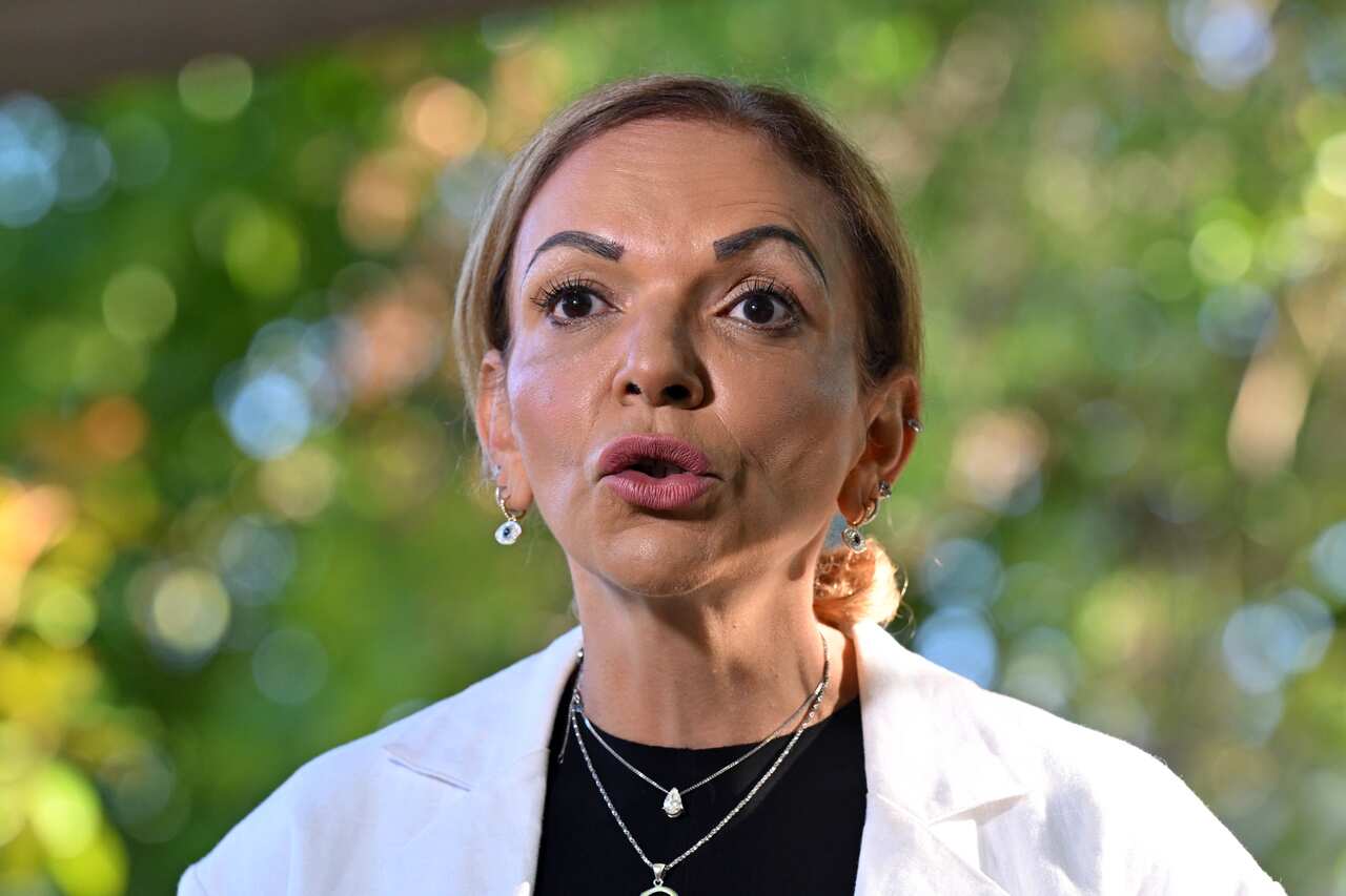 Anne Aly wearing a white shirt speaking in front of green trees.