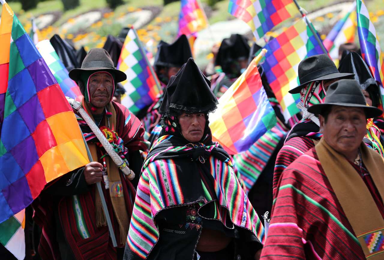 A group of people in traditional attire march, holding colourful flags.