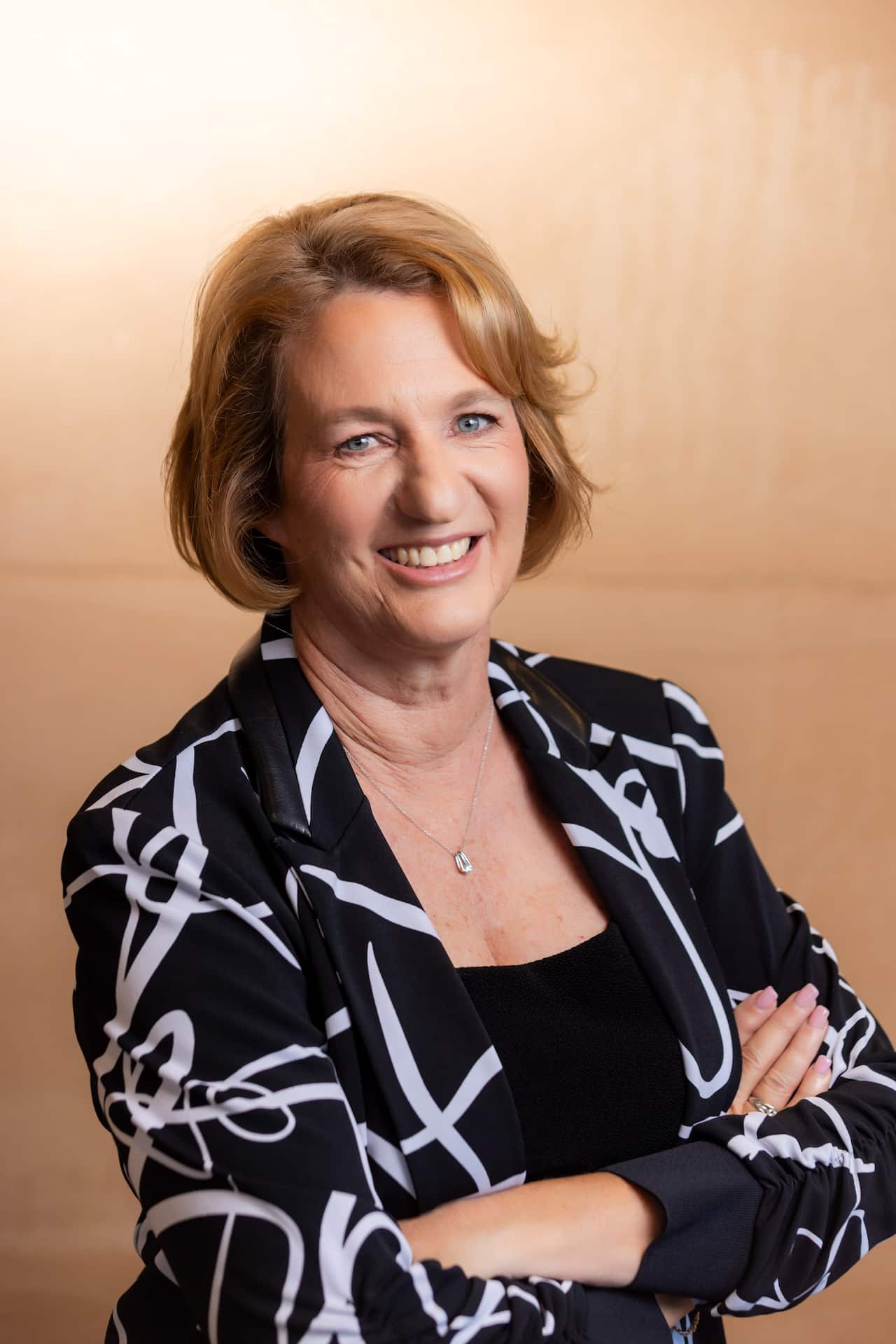 A woman in a black blazer with a white pattern smiles with her arms folded.