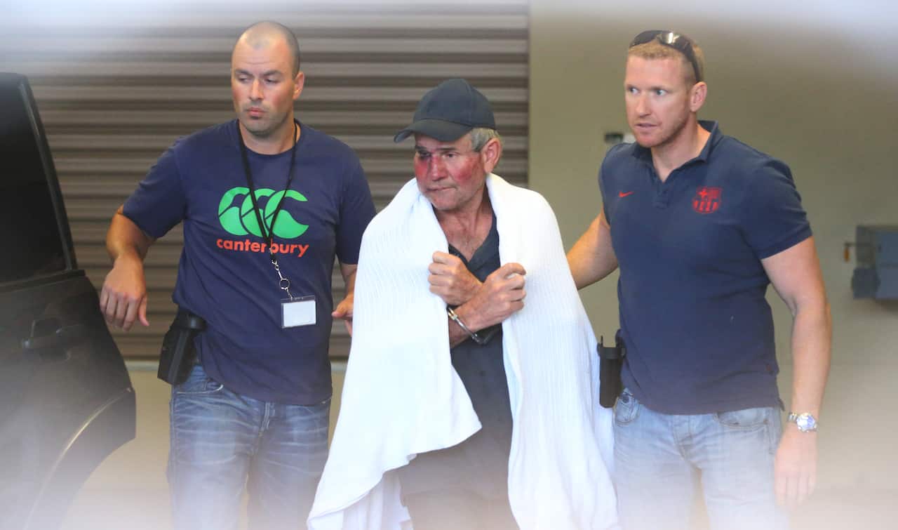 A man in a baseball cap with a towel draped over his shoulders is being escorted by two plainclothes police officers. He is handcuffed and appears to have a bruised face.