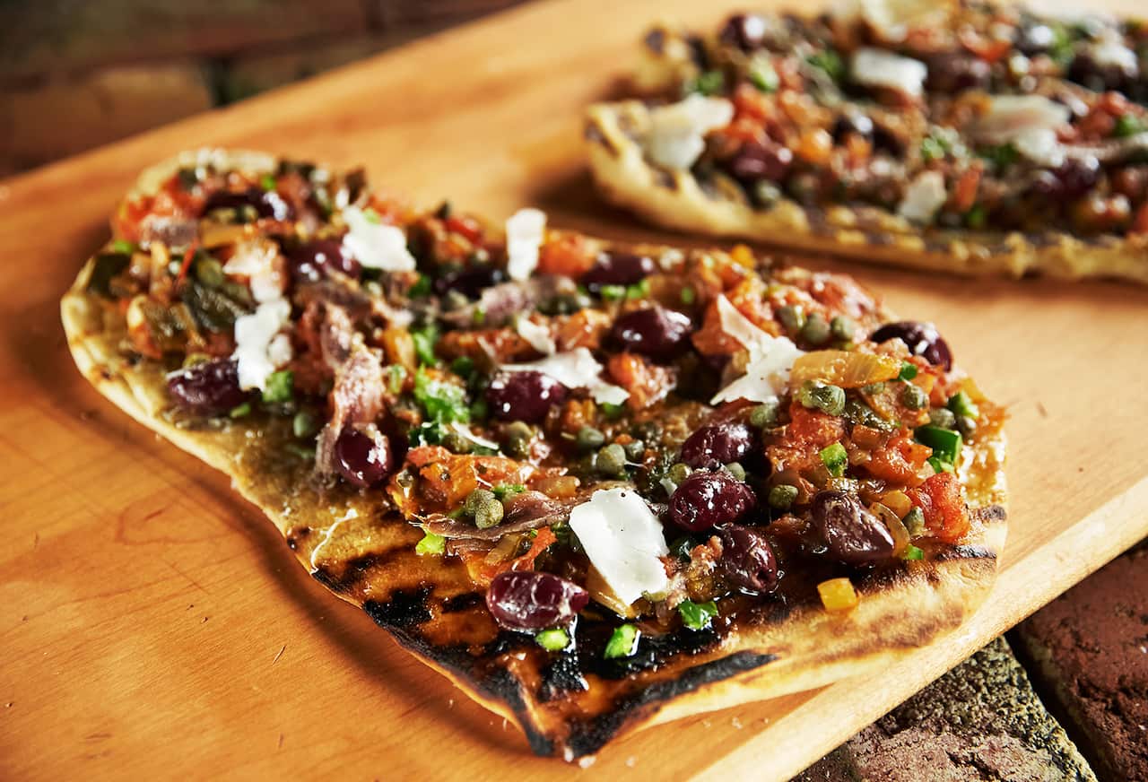 A roughly rectangular pizza with a chunky topping sits on a wooden board. Another pizza xan be seen behind it. 