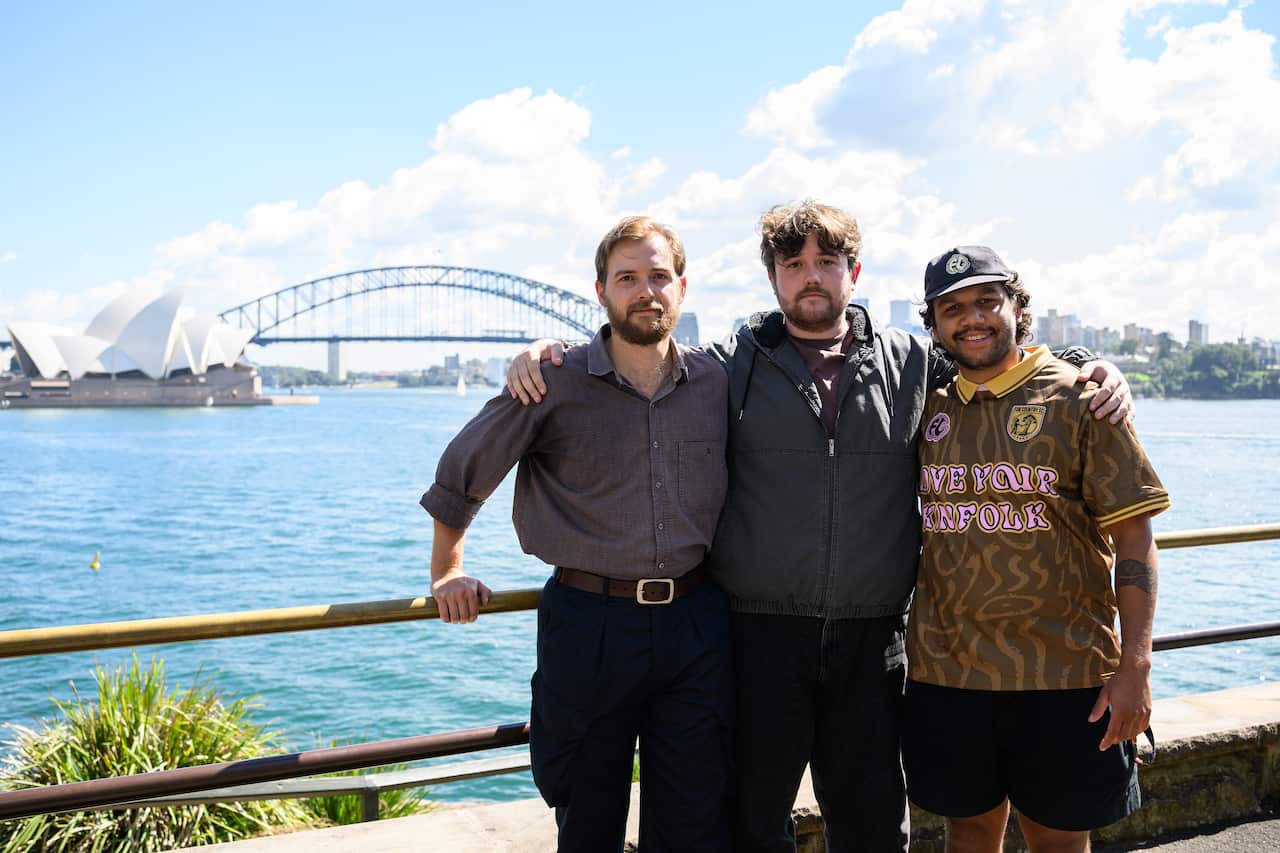 Three men stand together with their arms around each other, posing for a photo along a waterfront railing. In the background, the Sydney Opera House and the Sydney Harbour Bridge are visible under a bright, partly cloudy sky. The man on the left wears a brown button-down shirt and navy trousers, the man in the center wears a grey zip-up jacket, and the man on the right wears a patterned brown shirt with the words "LOVE YOUR KINFOLK" and a black baseball cap.