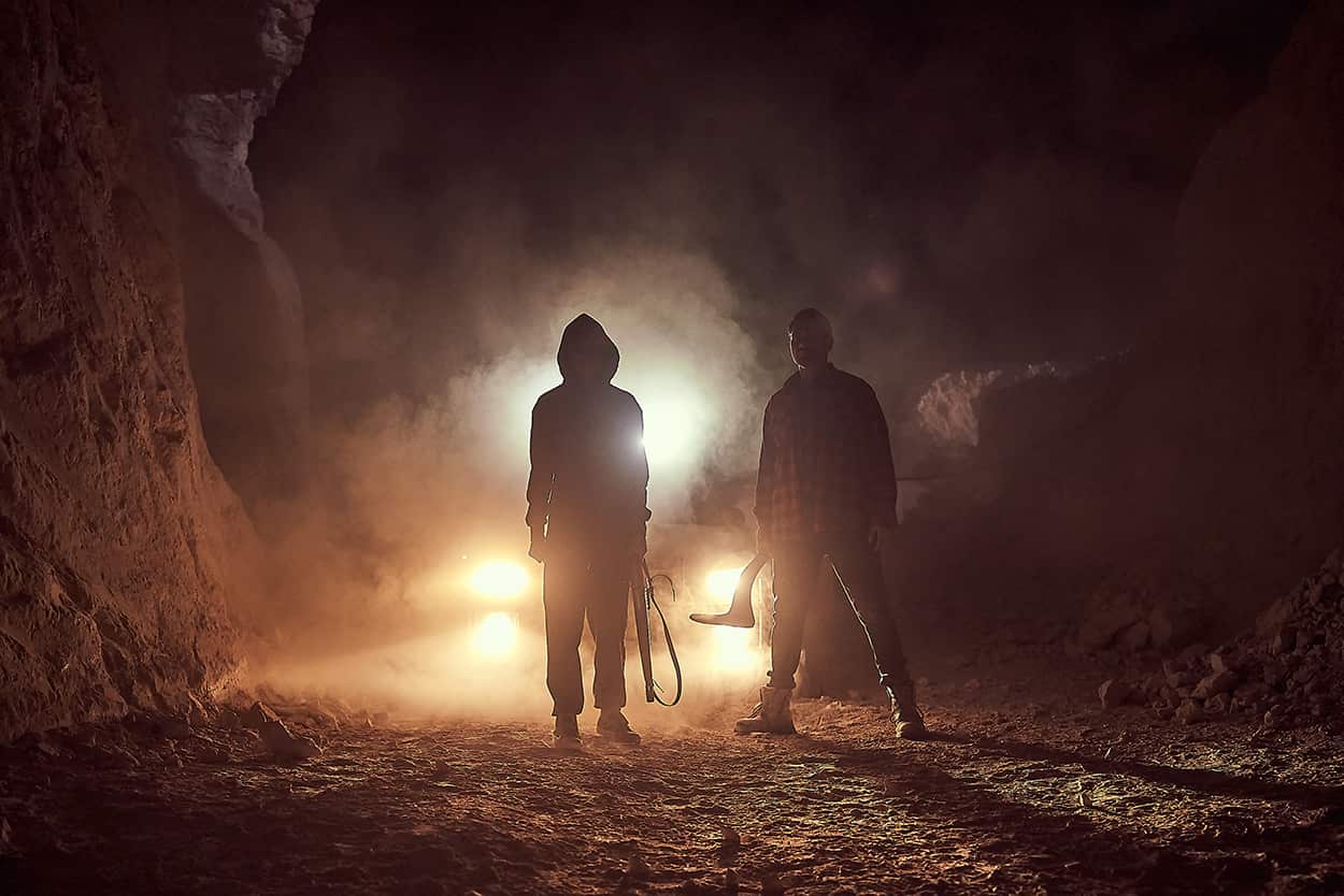 A young woman and a man stand in the outback at night, the headlights of a car behind them. They hold weapons.jpg
