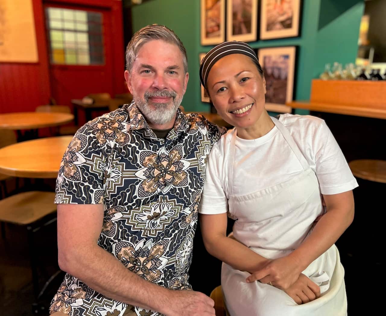 A man in a batik shirt sits hugging a woman in a white apron. 