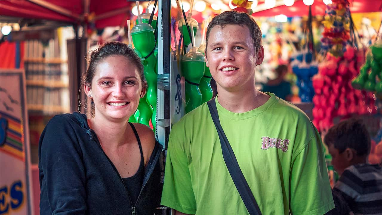 Two young people stand in front of a sideshow ally attractions. 