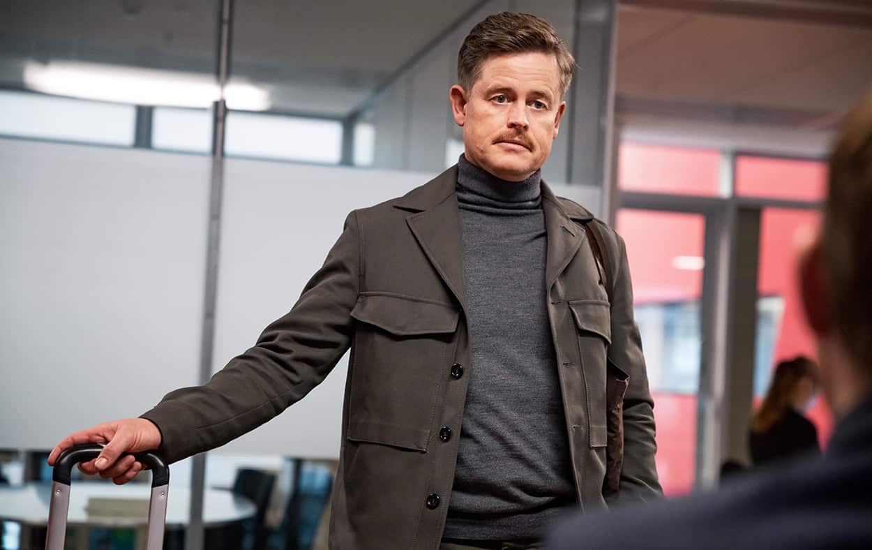 A man with brown hair and moustache looks down at a shadowed figure sitting in front of him.
