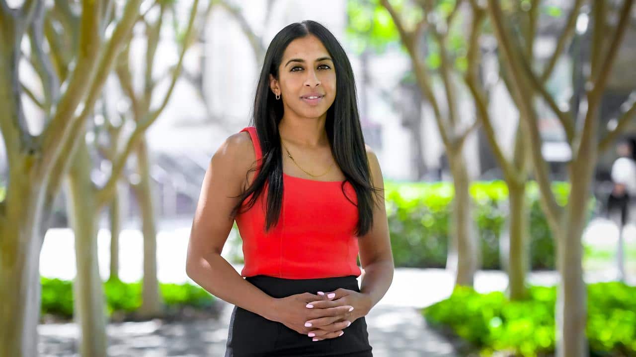 A woman with long dark hair and wearing a black dress and orange strappy stop stands with a neutral in a park. There are trees in the background