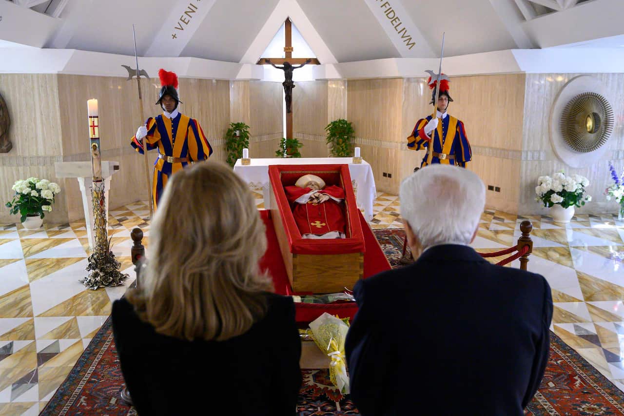 A man and a woman pay respects to late Pope Francis, whose body lies in a simple wooden coffin. Two Swiss Guards are standing on either side of the coffin.