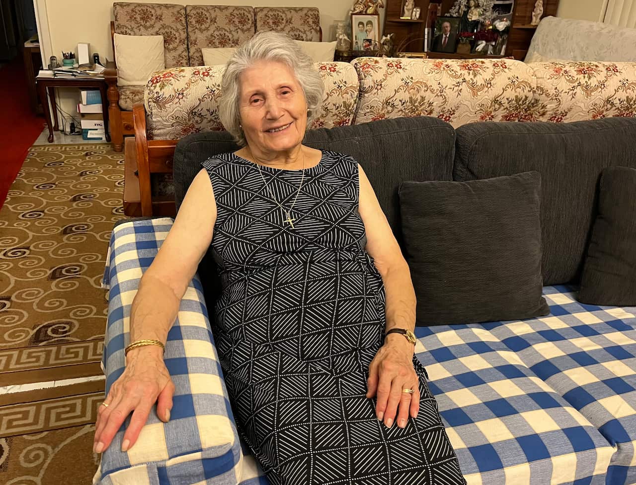 An elderly woman wearing a black dress smiles as she sits on a blue-and-white chequered sofa with black cushions in a living room.