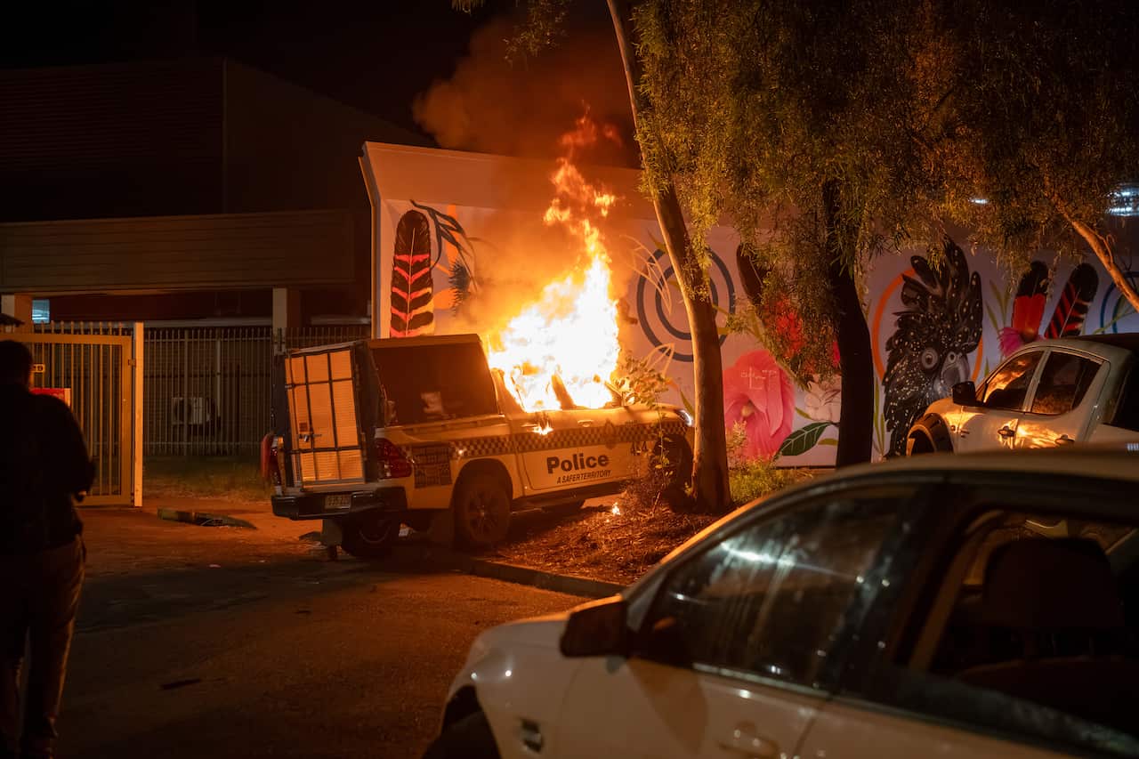 At nighttime, a police car driven onto a verge is engulfed in flames, one onlooker stands to the far left of the flame.