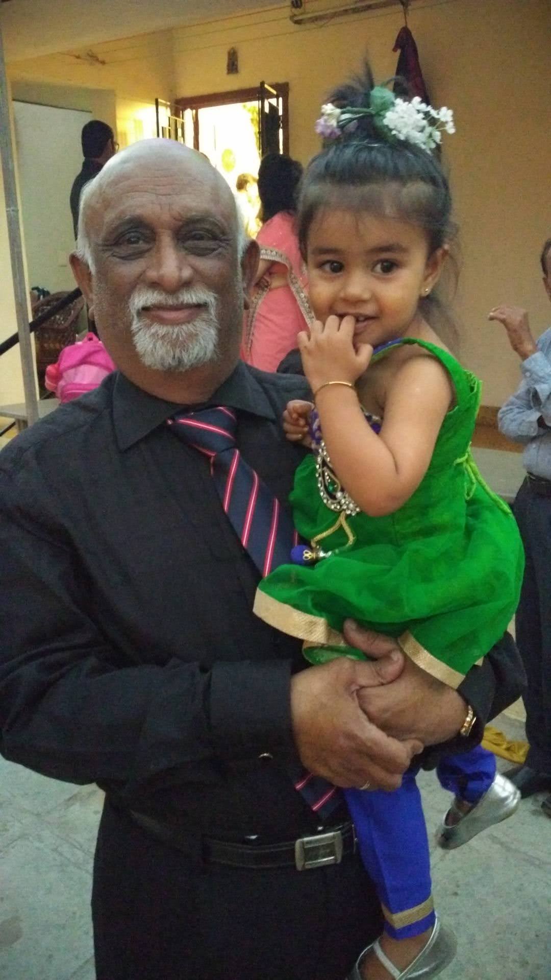 An elderly man in a black suit poses for a photo while holding a little girl wearing a green and blue dress.