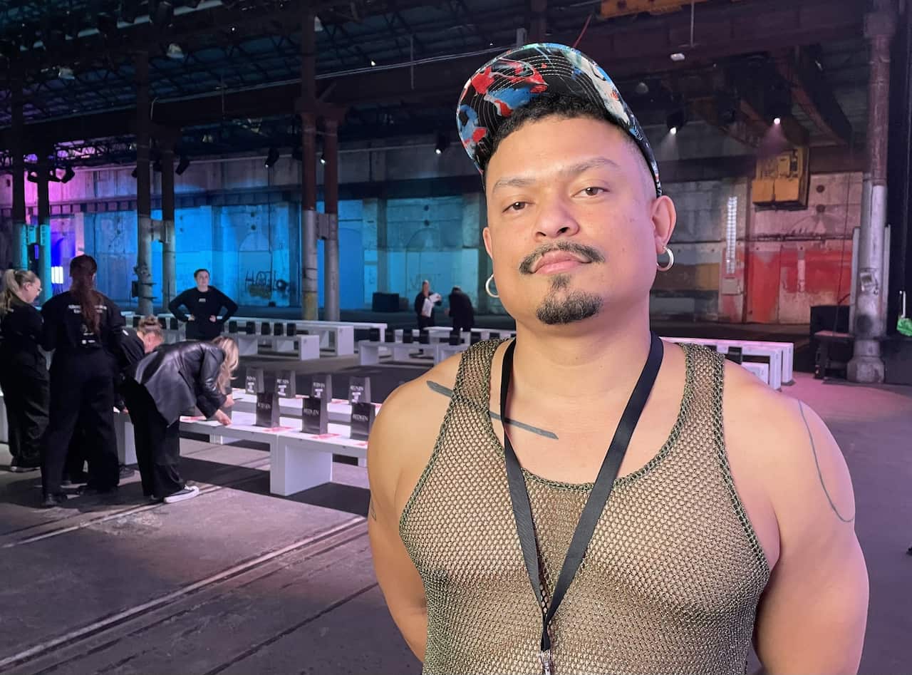 A man in a khaki mesh vest and a colourful baseball cap. He has a facial hair and is wearing silver hoop earrings