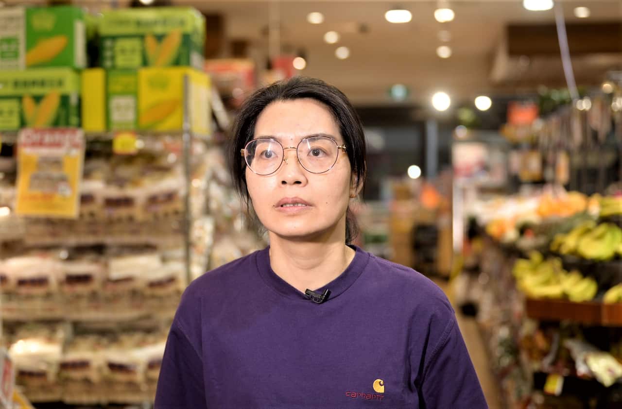 A woman wearing glasses and a purple jumper stands in the aisle of a supermarket.