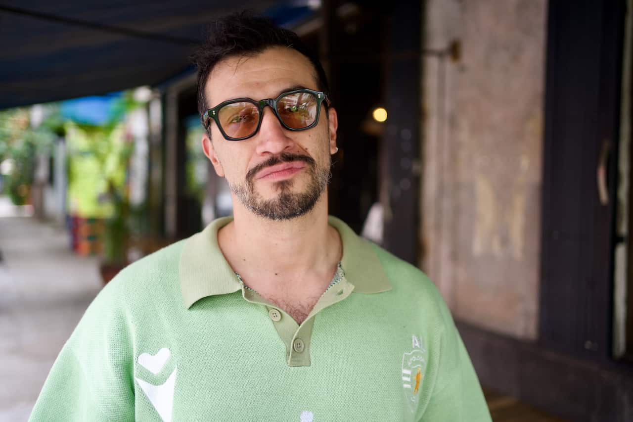 A close-up portrait of a man with dark hair, a beard, and a moustache, wearing black-rimmed sunglasses and a light green polo shirt, standing outdoors on a footpath.