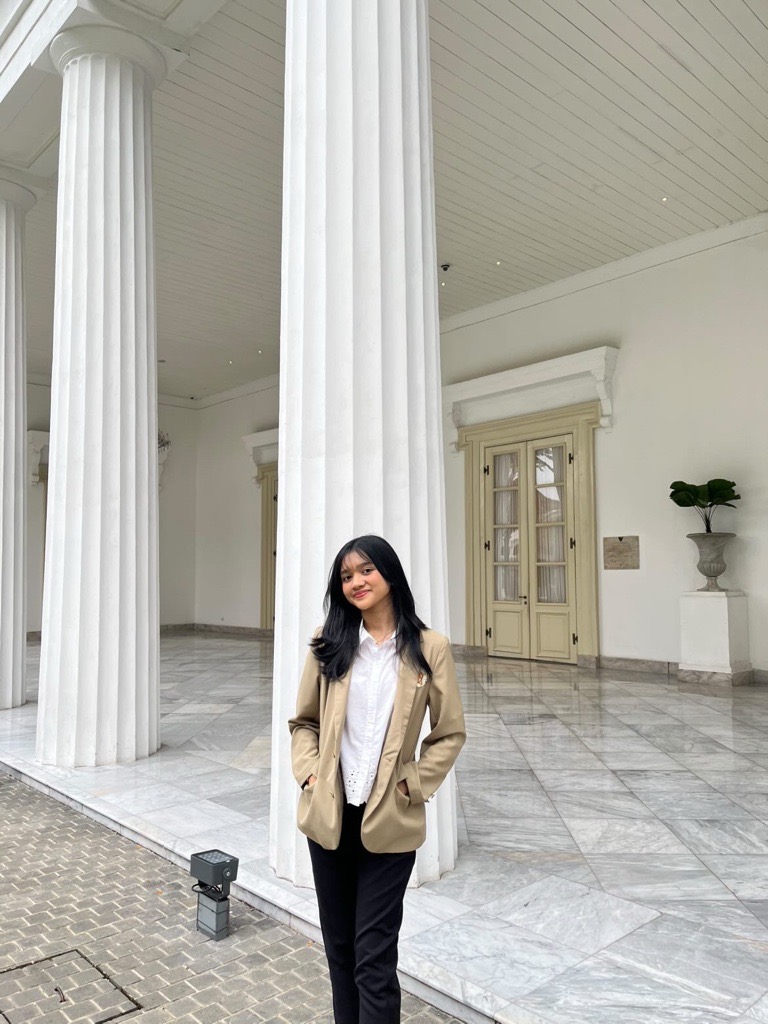 A young woman in a beige jacket stands outside a white building with tall pillars.