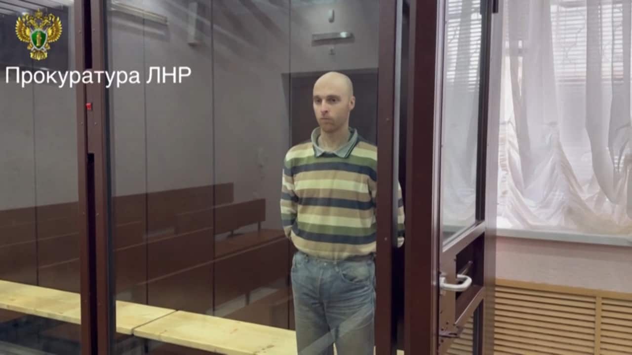 A man standing behind glass windows, wearing a striped shirt, blue jeans and with a despondent look on his face.