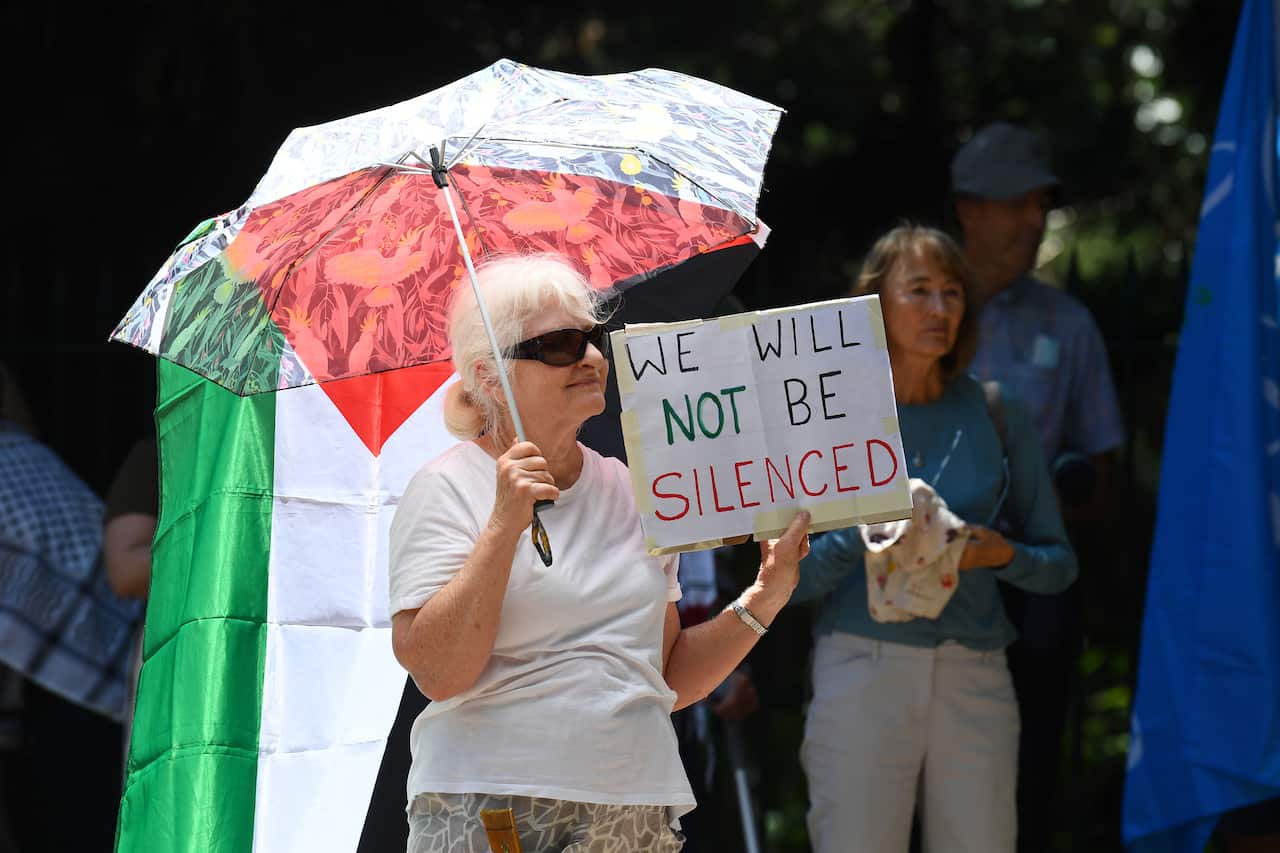 An old woman holds up a placard at a rally, she is also holding an umbrella over her head 