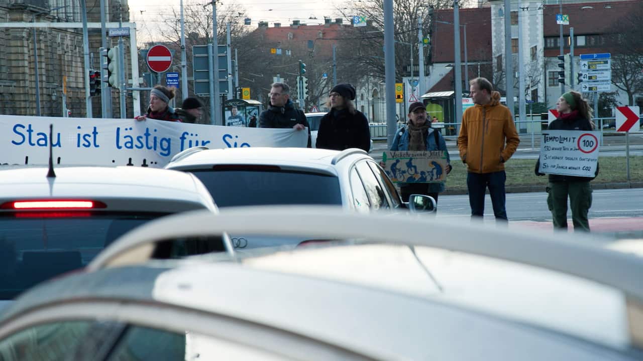 A group of men and women holding banners in the German language are standing in front of cars as they block traffic on the street