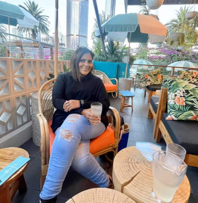 A woman wearing ripped blue jeans sitting on a wicker chair holding a drink 