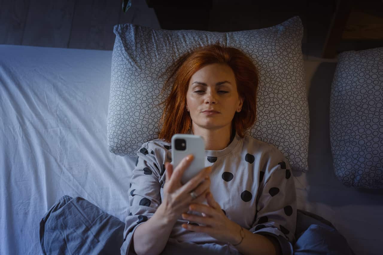 A woman with orange hair wearing a long sleeve grey shirt with blue dots, lying in bed holding up her phone.