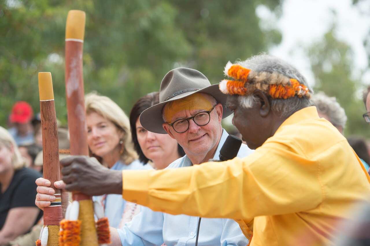 Anthony Albanese, wearing a light blue shirt and a hat and holding a didgeridoo, listens to a man wearing a yellow shirt and a coloured headband and holding a didgeridoo.  