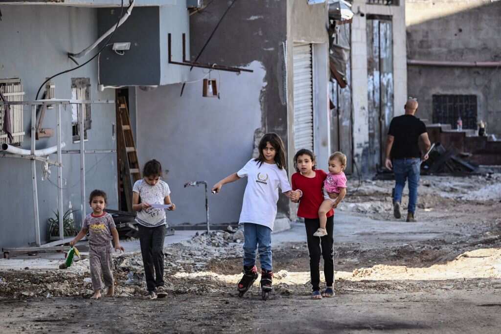 A row of children wander through the streets, which are full of rubble.