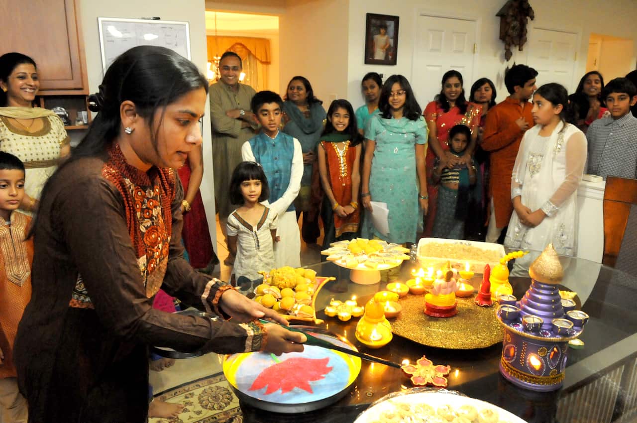 A woman lights a flower-shaped lamp placed on a table laden with traditional Indian sweets and earthen lamps in various shapes and sizes. She is surrounded by her family members indoors.