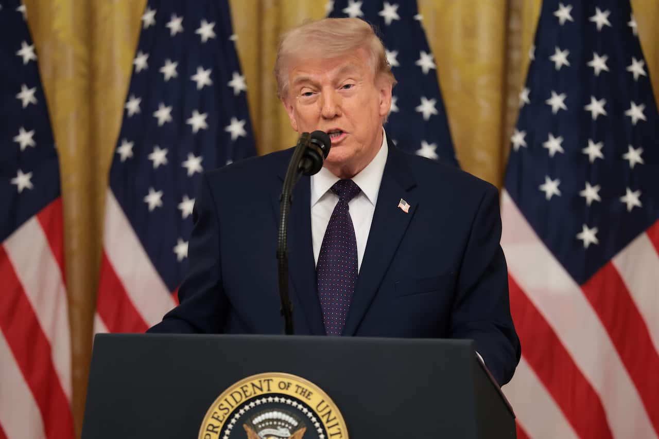 Wearing a dark suit and tie, Donald Trump speaks into a microphone at a podium with the US President Coat of Arms on the front. US flags hang behind him.