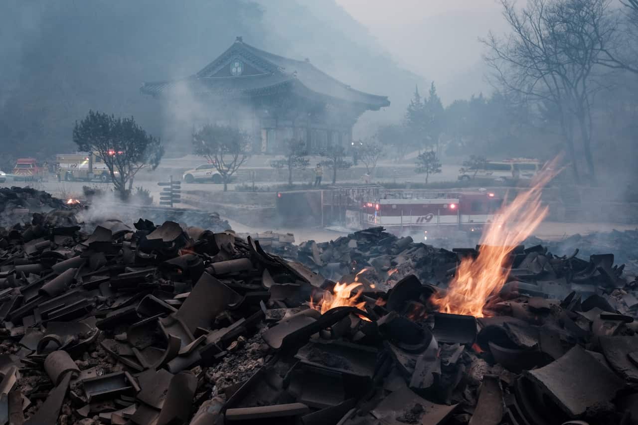Residual flames burn on top of wreckage as a temple sits in the background.